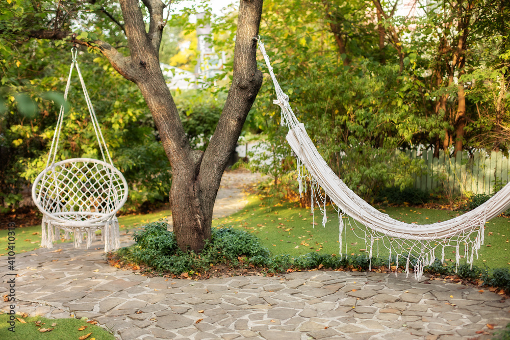 Comfortable hanging wicker white chair in summer garden. Cozy hygge place for weekend relax in garden. Hammock chair in boho style hanging on tree. Cozy exterior backyard. Concept of rest outdoor.
